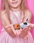 Two Guinea Pig toys in a childs hand with a pink background.